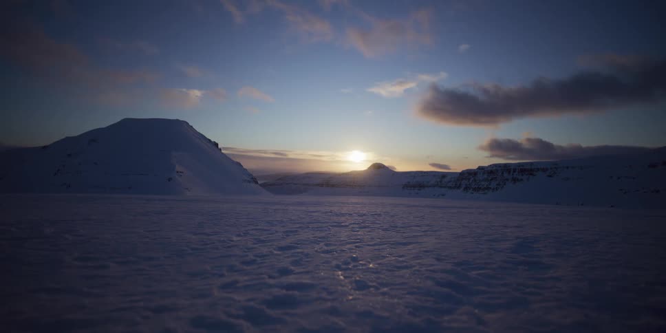 The setting sun casts long shadows across a vast, frozen valley surrounded by majestic snow-capped mountains