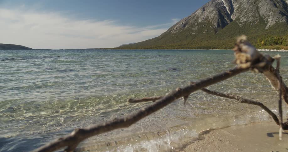 A tranquil scene shows gentle waves from a crystal clear lake lapping a sandy beach with a majestic mountain in the background