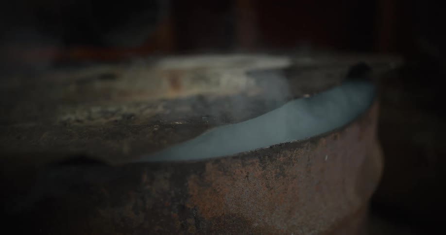 A close-up shot of thick white smoke billowing slowly from a rusty old metal barrel in a dark setting