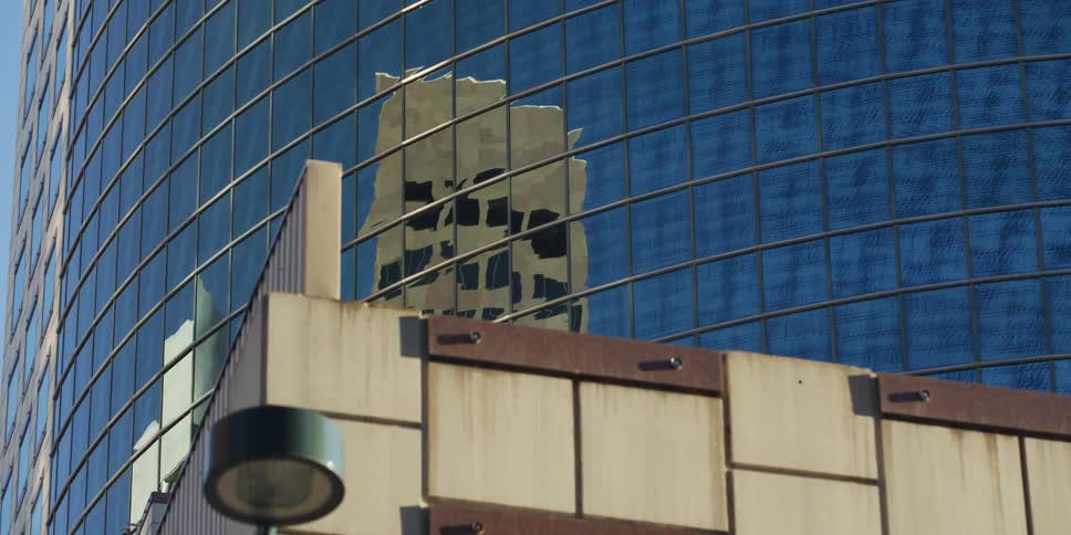 The distorted reflection of a city building is seen in the blue glass facade of a modern skyscraper