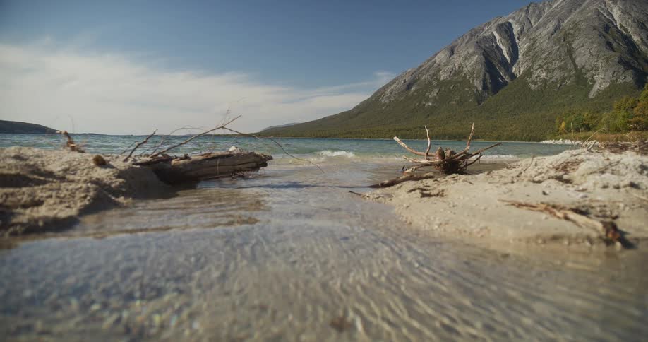 A clear stream flows across a sandy beach into a tranquil mountain lake on a sunny day
