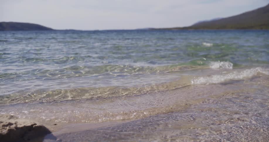 Crystal clear water gently laps onto a sandy beach in slow motion on a calm, sunny day