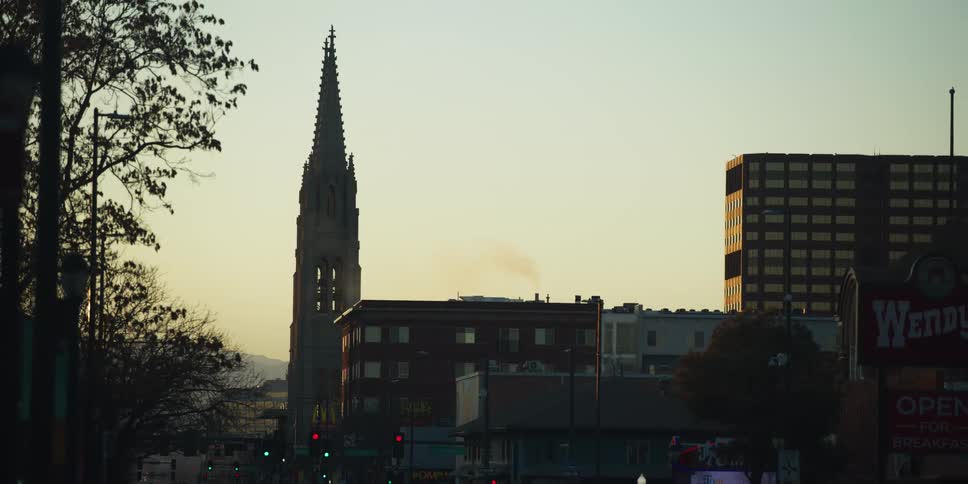 A tall church steeple towers over city buildings as traffic moves below during a hazy sunrise