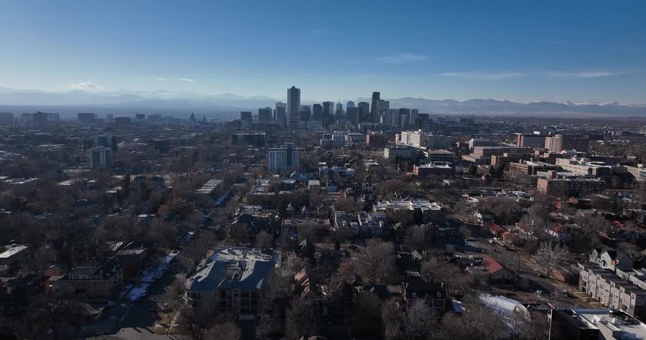 A slow aerial pullback reveals the expansive Denver skyline with the snow-capped Rocky Mountains in the background on a sunny day
