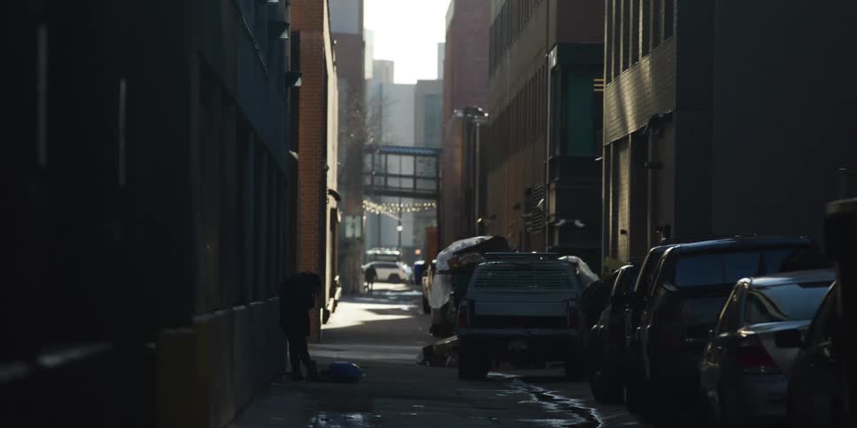 A homeless person is silhouetted in a dark city alley, sorting through their belongings on the ground