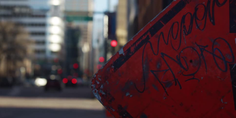 A weathered red street sign covered in graffiti sits in the foreground of a blurry urban city street with traffic