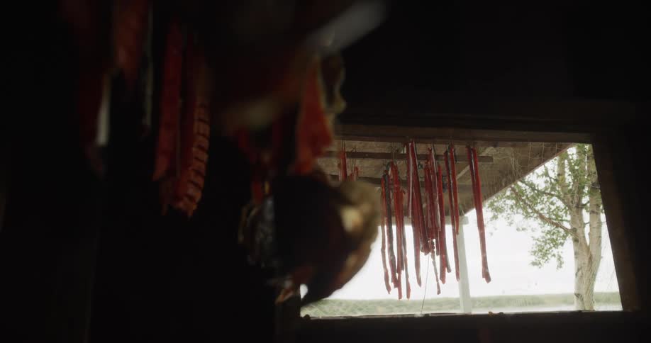 Strips of red salmon hang to dry in a dark smokehouse with a view of a tranquil river outside the window