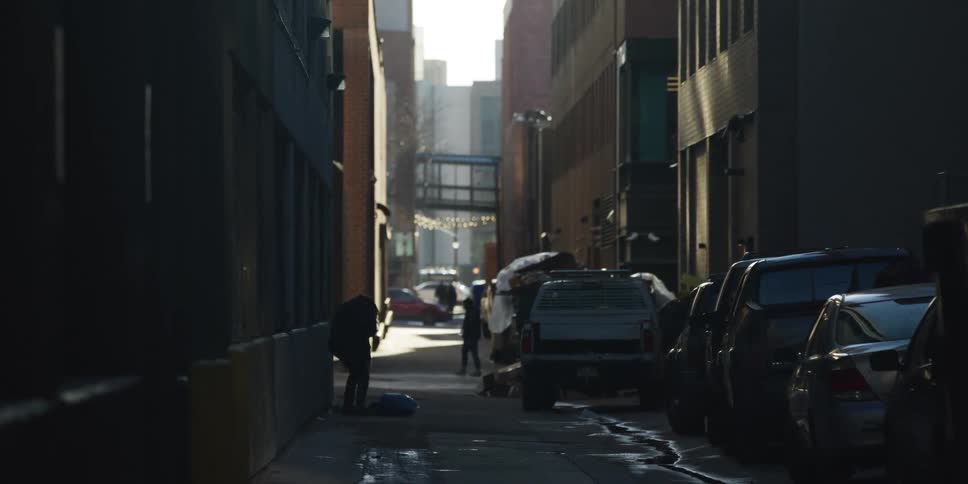 A silhouetted person rummages through their belongings in a dark, sunlit urban alleyway