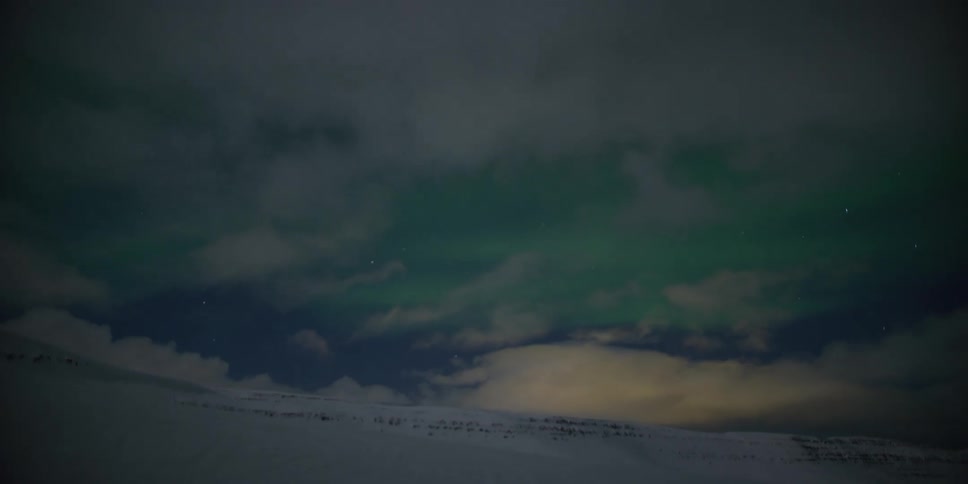 Green northern lights (aurora borealis) dance in a starry night sky above a snowy landscape as clouds pass by in this breathtaking time-lapse