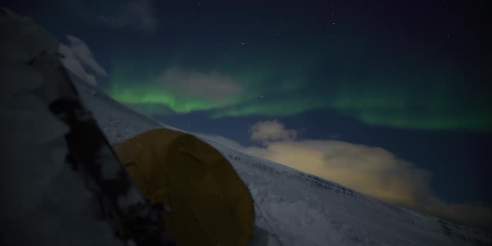 Time-lapse of the vibrant Northern Lights dancing in the starry night sky over a yellow tent pitched on a snowy mountain
