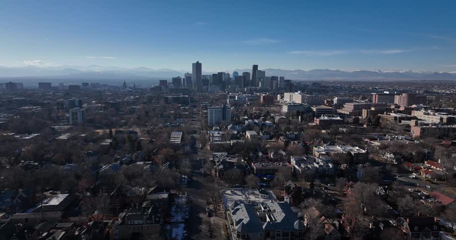 A slow aerial pullback shot reveals the Denver, Colorado skyline with the majestic Rocky Mountains in the background on a clear, sunny day