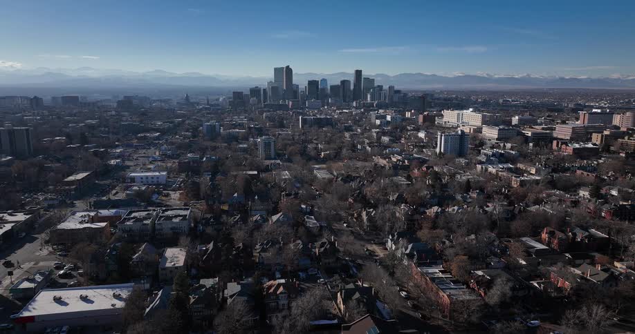 A slow, wide aerial pullback shot reveals the Denver, Colorado skyline with the Rocky Mountains in the distant background on a clear, sunny day