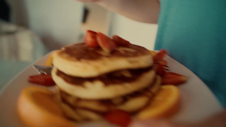 A person brings a plate of pancakes garnished with strawberries and oranges to serve as breakfast in bed