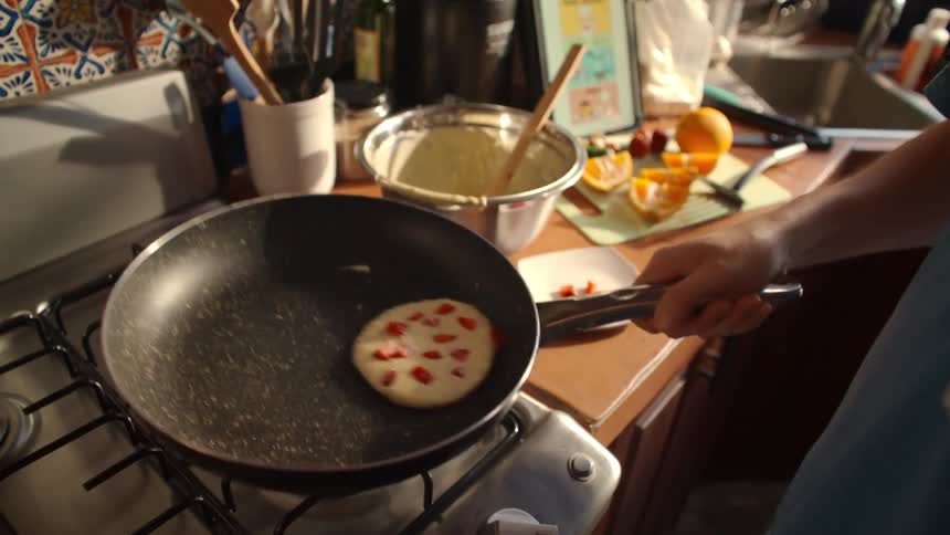 A person skillfully flips a fresh strawberry pancake in a pan over a gas stove in slow motion