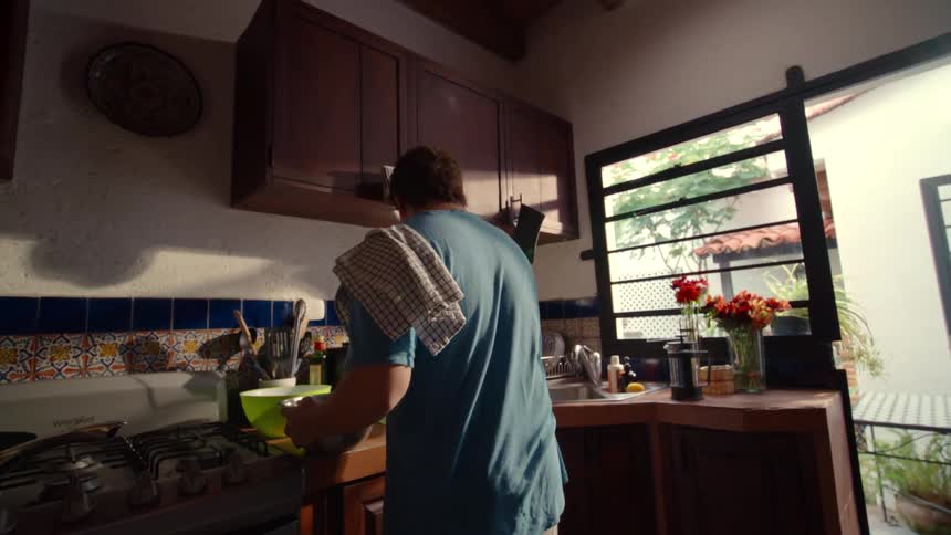 A man follows a recipe on his laptop while cooking in a sunlit, rustic-style kitchen