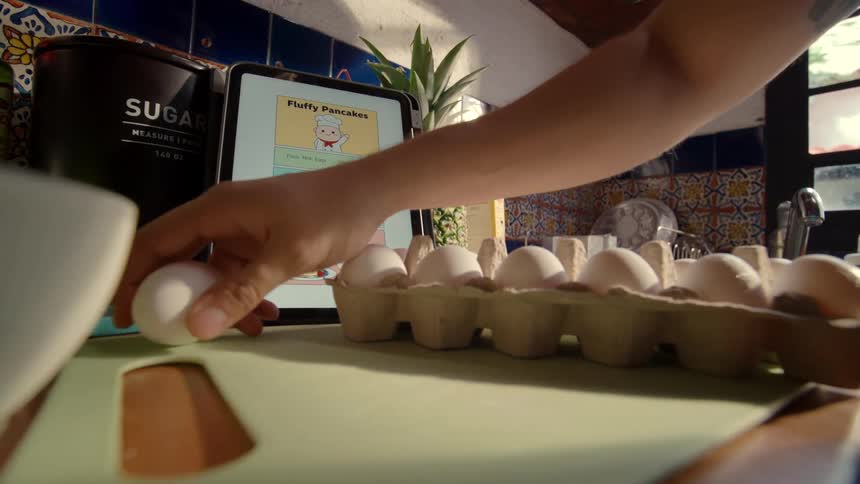 Following a cute illustrated recipe on a tablet, a person cracks an egg into a bowl to make fluffy pancakes for breakfast
