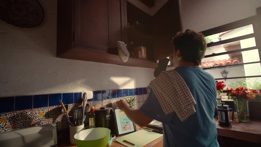 Young man gathers ingredients from a cabinet while following a recipe on a tablet in a sunny kitchen