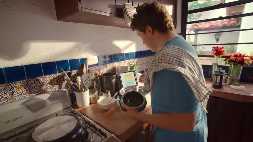 A young man follows a recipe on a tablet, measuring ingredients into a mixing bowl in a sunny, rustic-style kitchen