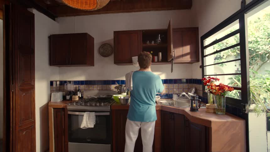 A man enters a rustic, sunlit kitchen to prepare food, washing his hands and fruit before getting supplies from a cabinet
