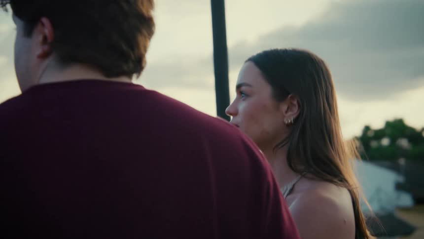 A man drinks a beer while talking to a woman on a rooftop during a beautiful sunset