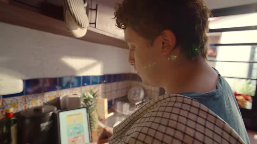 Over the shoulder shot of a man whisking eggs in a bowl, following an online recipe for pancakes on his tablet in a cozy, sunlit kitchen