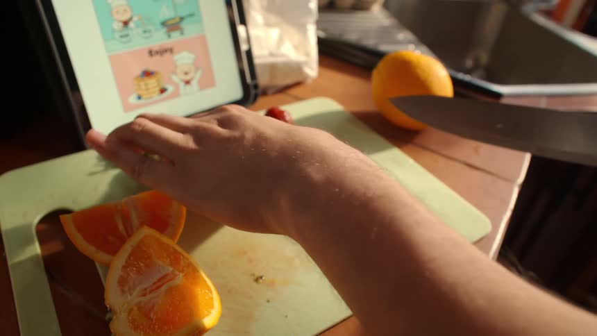 A person follows an online recipe while slicing a fresh orange on a cutting board in a sunlit kitchen