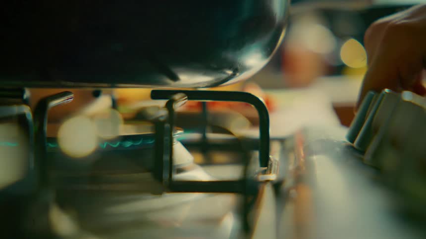 A macro shot of a hand turning the knob on a gas stove, igniting the blue flame to heat a pot