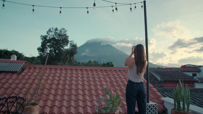 A woman on a rooftop terrace takes a photo of a volcano with her phone as a man joins her with a glass of wine