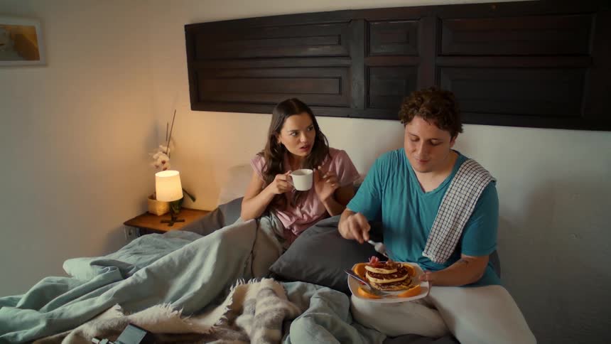 A happy young couple enjoys a romantic breakfast in bed, with the man playfully feeding his partner a bite of pancake