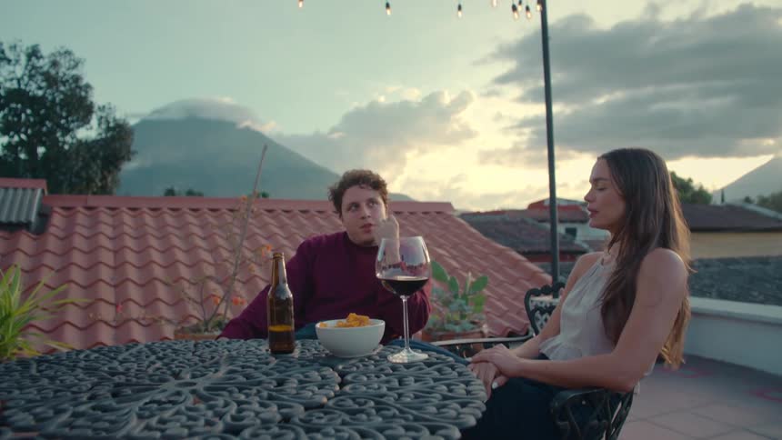 A happy young couple enjoys drinks and conversation on a scenic rooftop patio at dusk with a volcano view