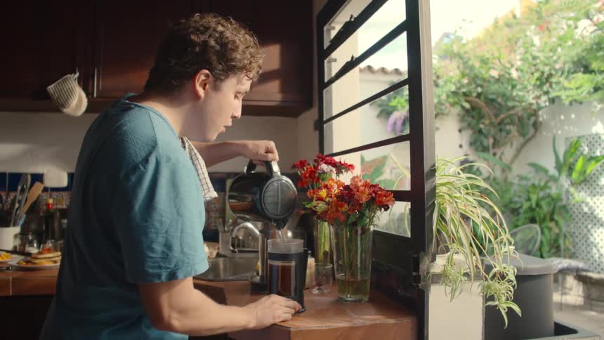 A man in a sun-drenched kitchen pours hot water from a kettle into a French press to brew coffee, with a vase of colorful flowers on the counter next to him