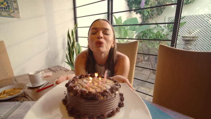 First-person perspective of surprising a young woman with a birthday cake while she studies, who then happily sings and blows out the candles