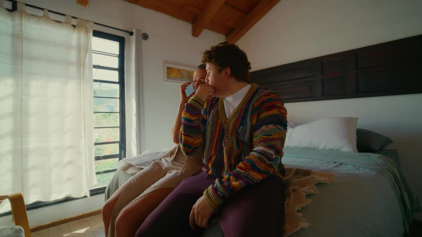 A young woman laughs while talking on the phone in a sunlit bedroom as her boyfriend sits beside her and whispers in her ear
