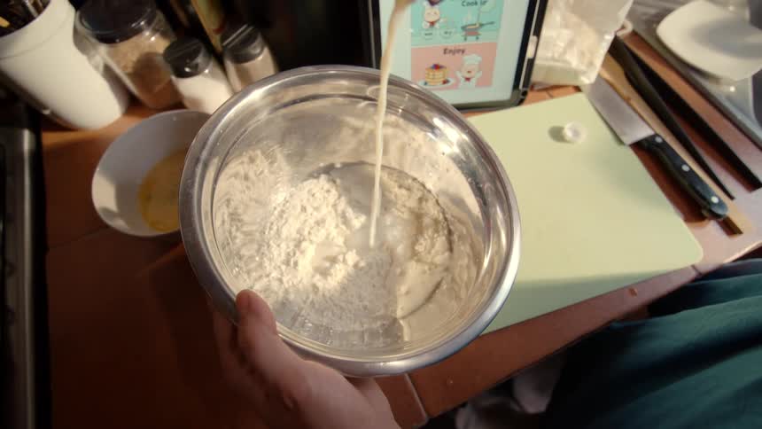A first-person perspective in slow motion shows a person pouring milk into a stainless steel bowl of flour while following a recipe on a tablet