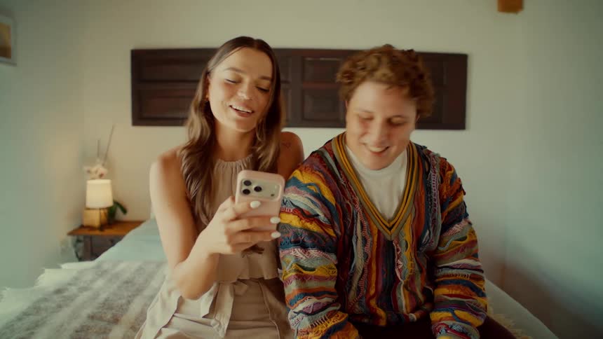 A playful young man and woman have a fun conversation and laugh while using a smartphone together on their bed