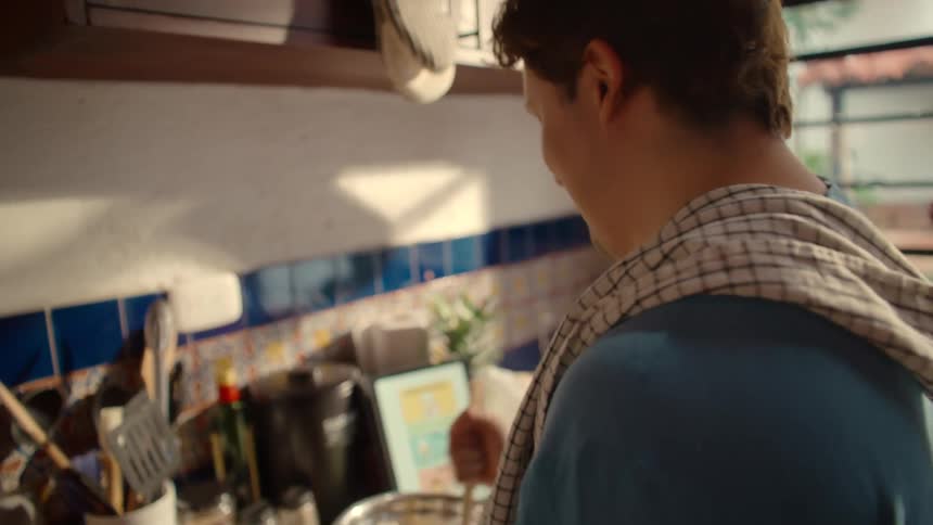 A young man enjoys a relaxing morning in a cozy, sunlit kitchen while mixing pancake batter and following an online recipe on a tablet