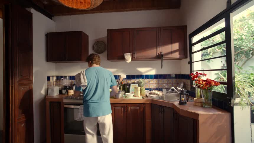 Rear view of a man cooking a meal in a cozy, sunlit kitchen with an open window to a garden
