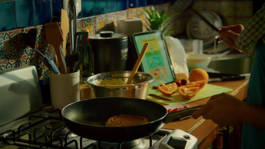 A person cooks breakfast in a cozy kitchen, flipping a pancake with berries in slow motion while following a recipe on a tablet