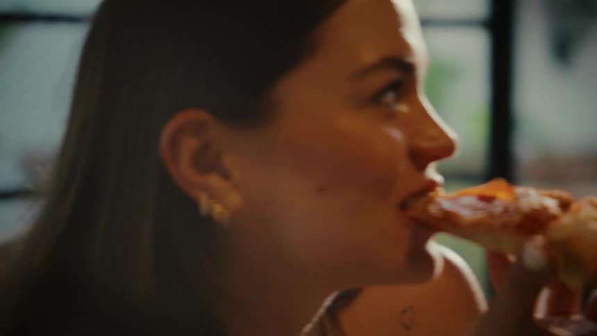 A close-up slow-motion shot of a beautiful young woman sensually taking a bite of a delicious slice of pizza