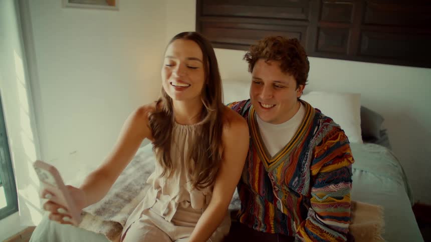 A happy young couple smiles and waves while having a fun video call on a smartphone in their cozy bedroom