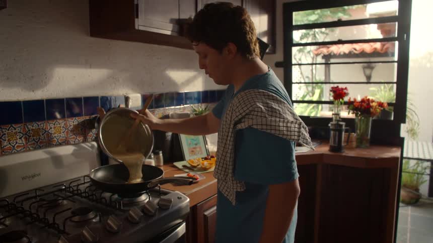 Young man in a sunlit kitchen follows a recipe on a tablet and pours pancake batter into a hot pan in slow motion