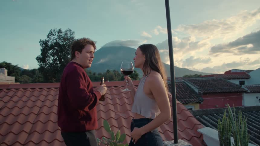 A happy young couple laughs and enjoys drinks on a scenic rooftop patio overlooking a town with a volcano in the background