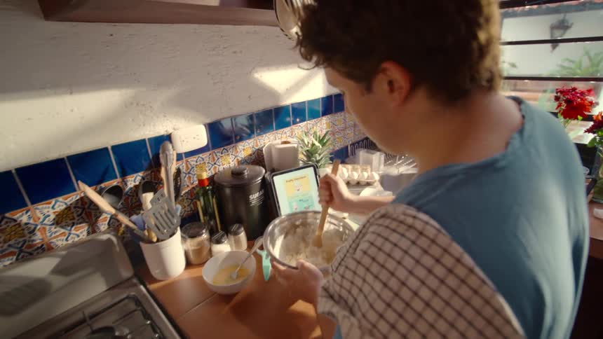 Man follows a digital recipe on his tablet, carefully mixing ingredients for a homemade meal in a warm, sunlit kitchen