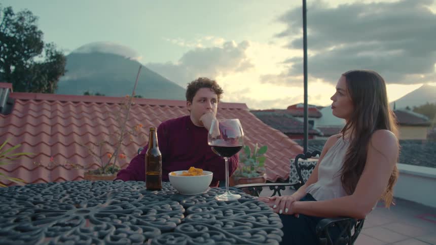 A young couple relaxes with wine and beer on a scenic rooftop patio, enjoying a conversation as the sun sets behind distant volcanoes