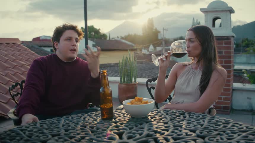 A young man and woman have a relaxed conversation while enjoying drinks on a rooftop patio with a view of the city and mountains during a beautiful sunset