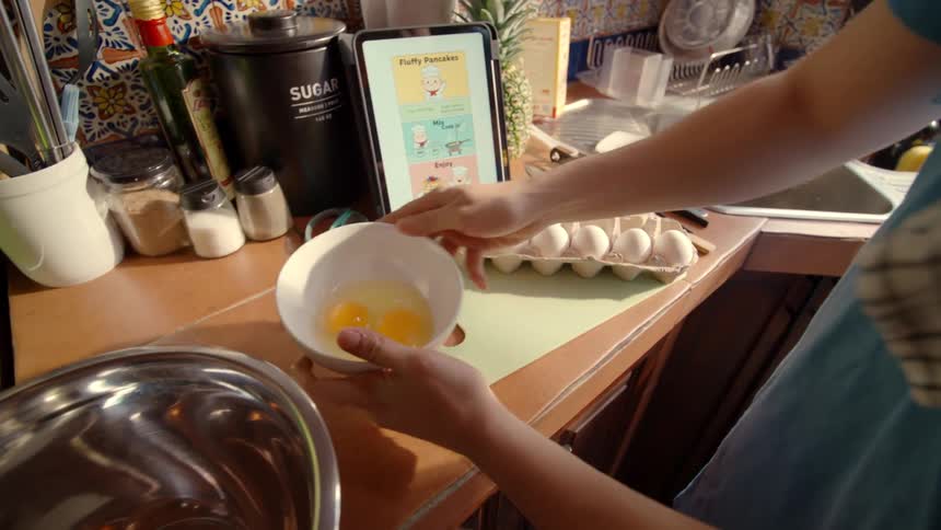 A person follows a digital recipe on a tablet to make fluffy pancakes, cracking and whisking eggs in a sunlit kitchen