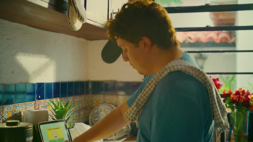 A man enjoys a taste of his cooking while following a pancake recipe on a tablet in a sunlit, rustic kitchen
