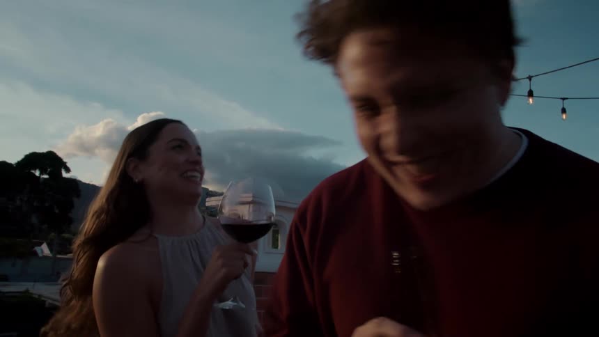 A happy young couple drinks, talks, and laughs on a rooftop patio at golden hour in slow motion