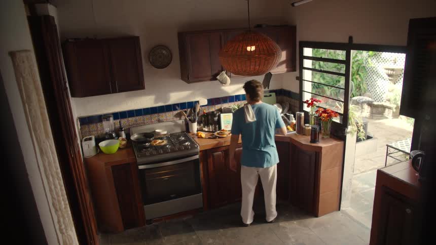 A man enjoys his morning routine, cooking and dancing in a cozy, sunlit kitchen as seen from a high angle