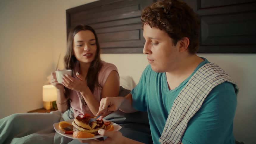 A loving man feeds his smiling girlfriend pancakes with fresh fruit for breakfast in bed